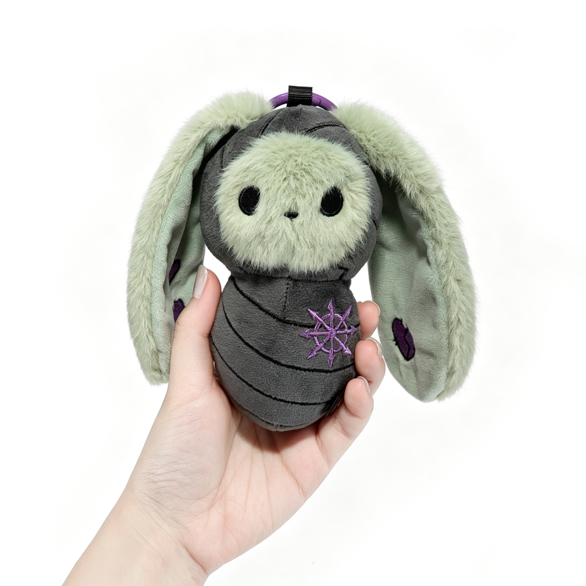 A hand holds a Bunnculus Plush Keychain: a green rabbit plush with stripes and purple star embroidery.